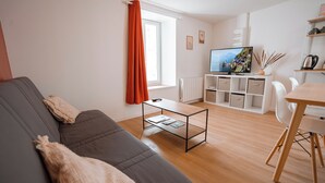 A modern living room with a grey sofa, a coffee table, and a dining area with a wooden table and white chairs. - Charming T2 in the center of Saint-Astier (Saint-Astier)