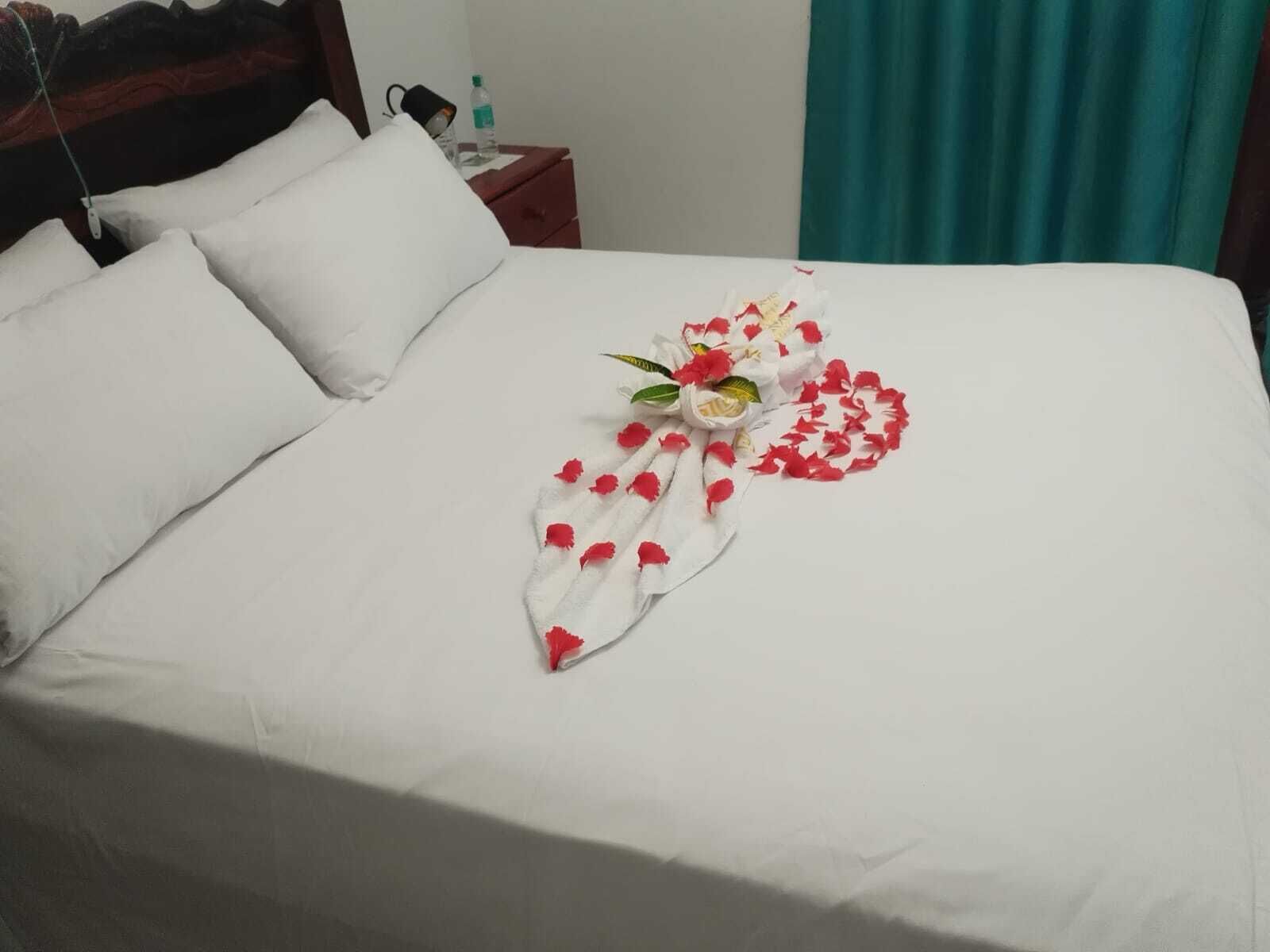 A neatly made bed with white linens, a pillow, and a decorative arrangement of red flower petals and a white cloth.