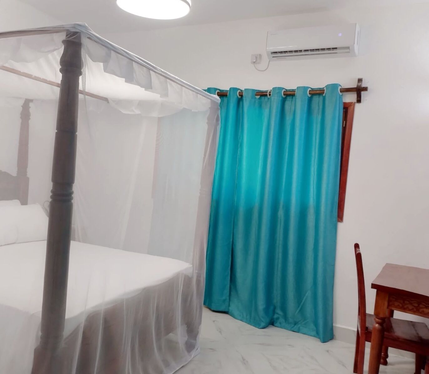 Une chambre avec un lit recouvert d’une moustiquaire, un rideau de couleur turquoise, une chaise en bois et un climatiseur fixé au mur.