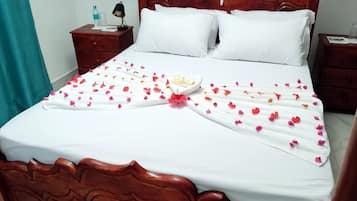 Una cama bien tendida con una colcha blanca, dos almohadas y una toalla decorativa con pétalos de flores.