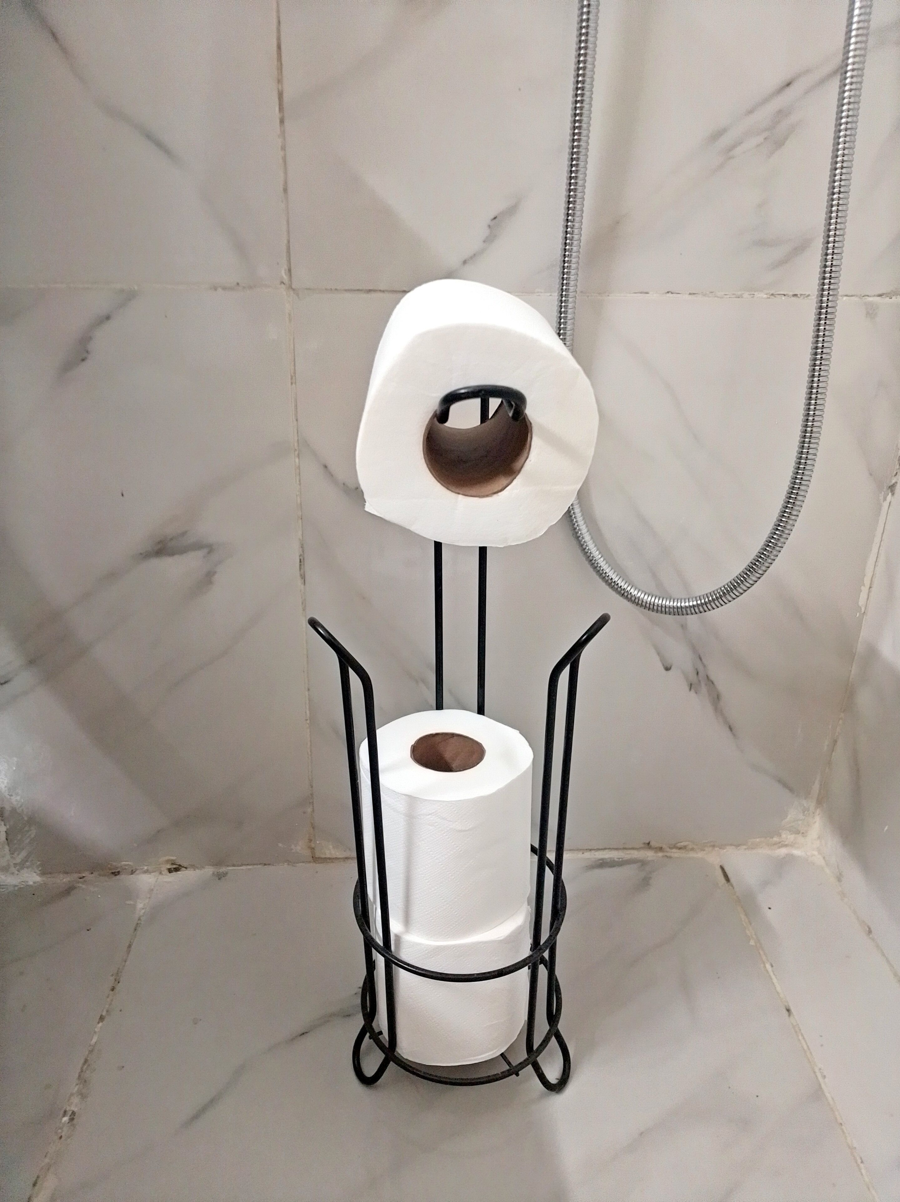 A toilet paper holder in a bathroom with marble walls and a shower hose.