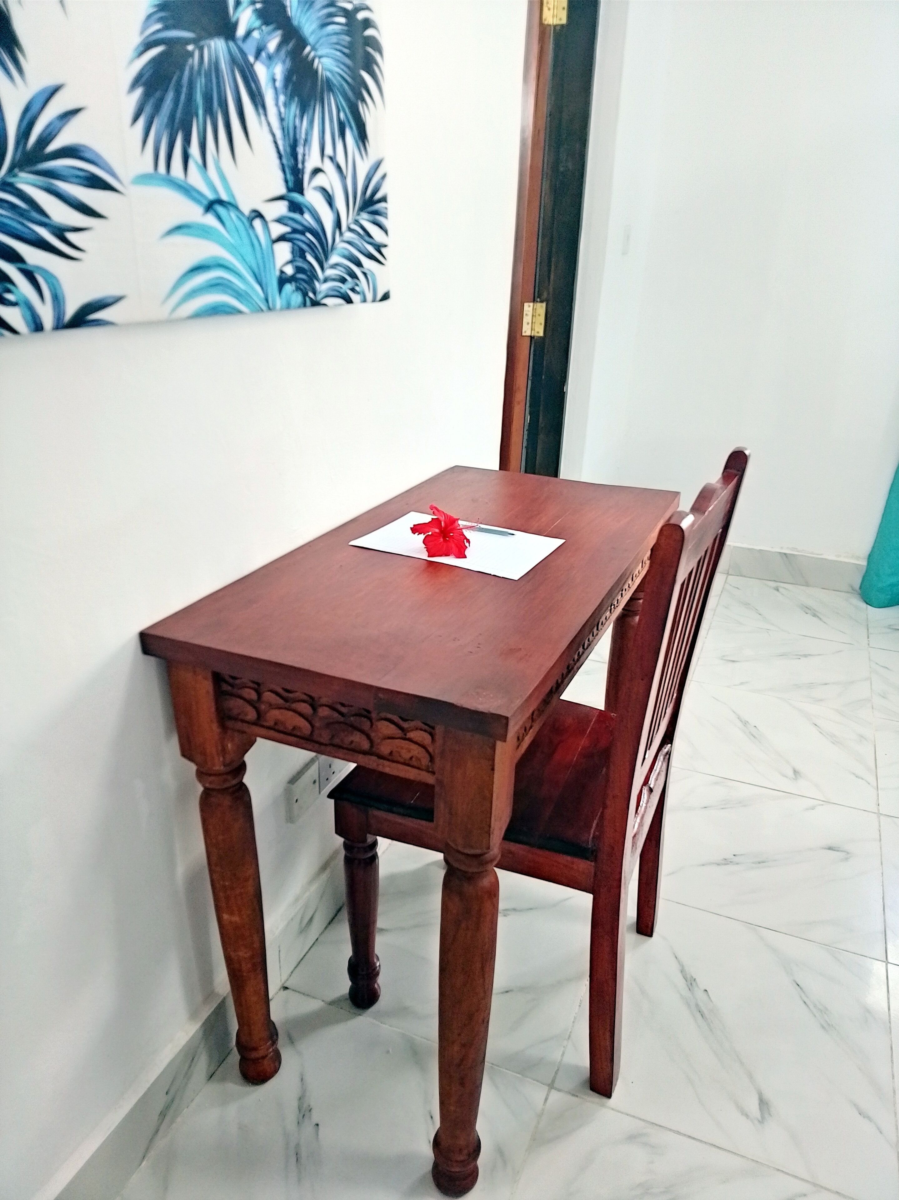 A wooden dining table with a red flower centerpiece, a wooden chair, and a wall with a blue and white tropical print.