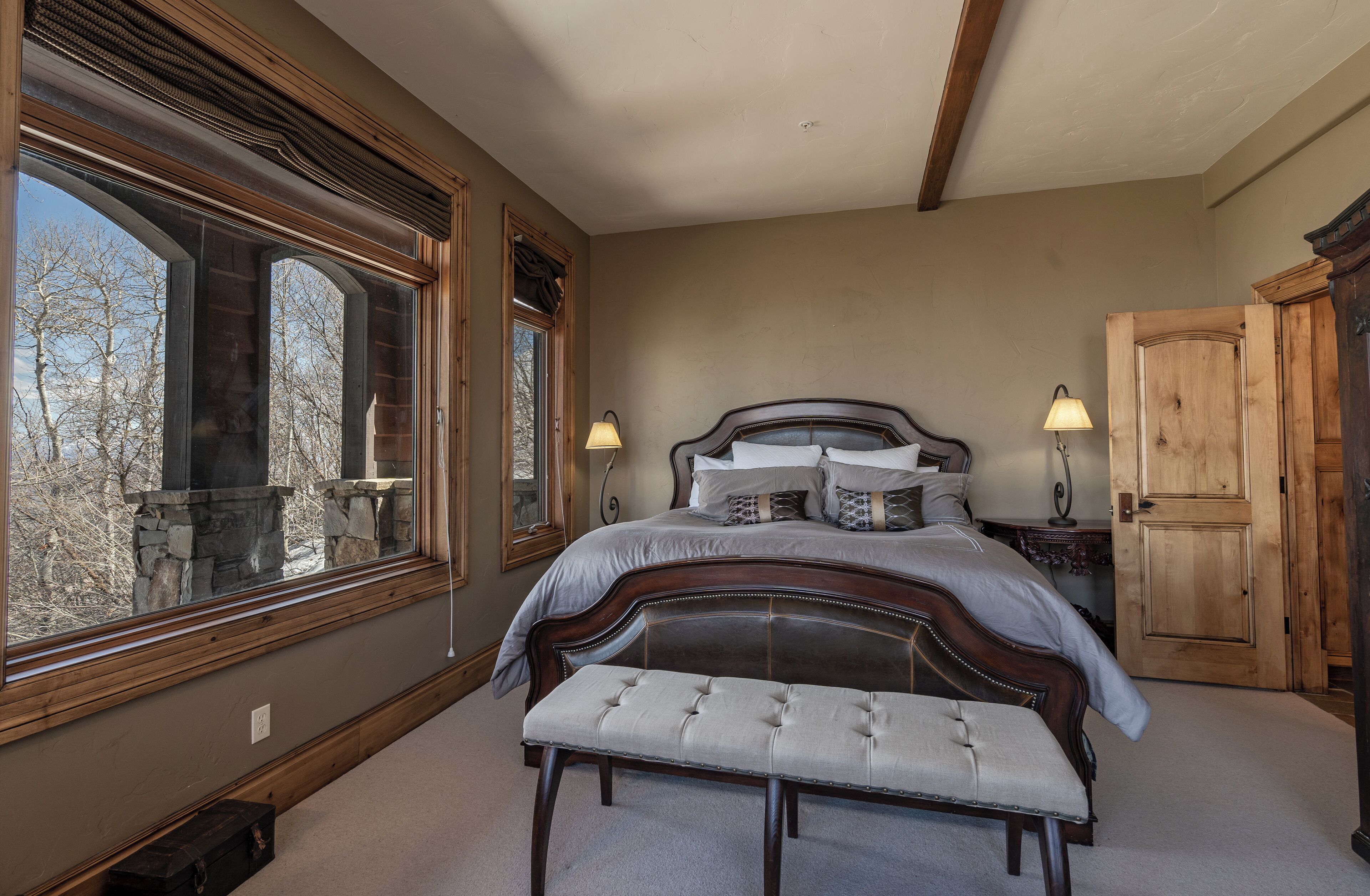 A bedroom with a large window, a wooden headboard, a bench, and two bedside lamps.