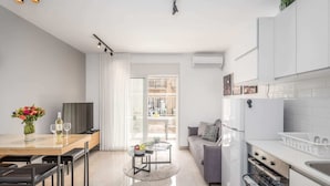Une cuisine moderne avec des meubles blancs, un coin repas comprenant une table en bois et des tabourets de bar noirs, un canapé gris et une petite table ronde sur laquelle sont posés des fruits et un magazine.