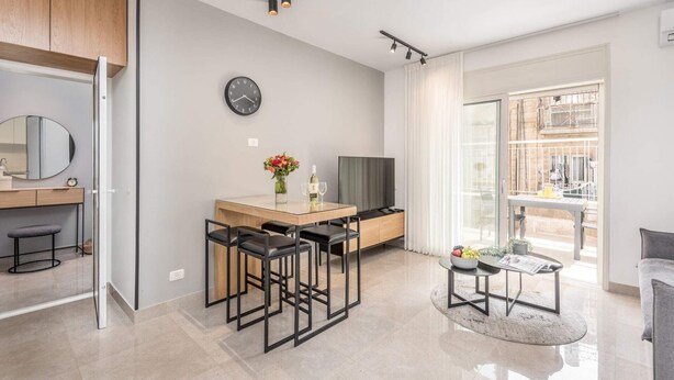 Un salon moderne avec une table à manger en bois, des chaises noires, un téléviseur et une grande fenêtre donnant sur des immeubles.