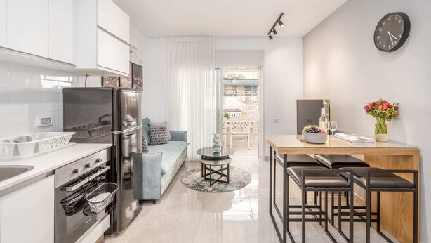 A modern kitchen with a refrigerator, oven, and sink. There is a dining area with a table and chairs, and a living area with a sofa and a coffee table. A clock is visible on the wall. - Machane Yehuda 4 - One Bedroom Apartment, Sleeps 4 (Jerusalem)