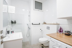 Un baño moderno con lavamanos, inodoro y ducha blancos. Hay una encimera de madera con una plancha y una cesta.