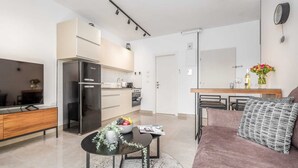 Un salon moderne comprenant un canapé, une table basse avec un saladier de fruits, un téléviseur et un coin cuisine avec un réfrigérateur et des placards.