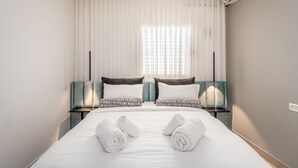 A hotel room with a large bed, two bedside tables, a nightstand, a window with sheer curtains, and an air conditioning unit. - Machane Yehuda 8 - One Bedroom Apartment, Sleeps 4 (Jerusalem)