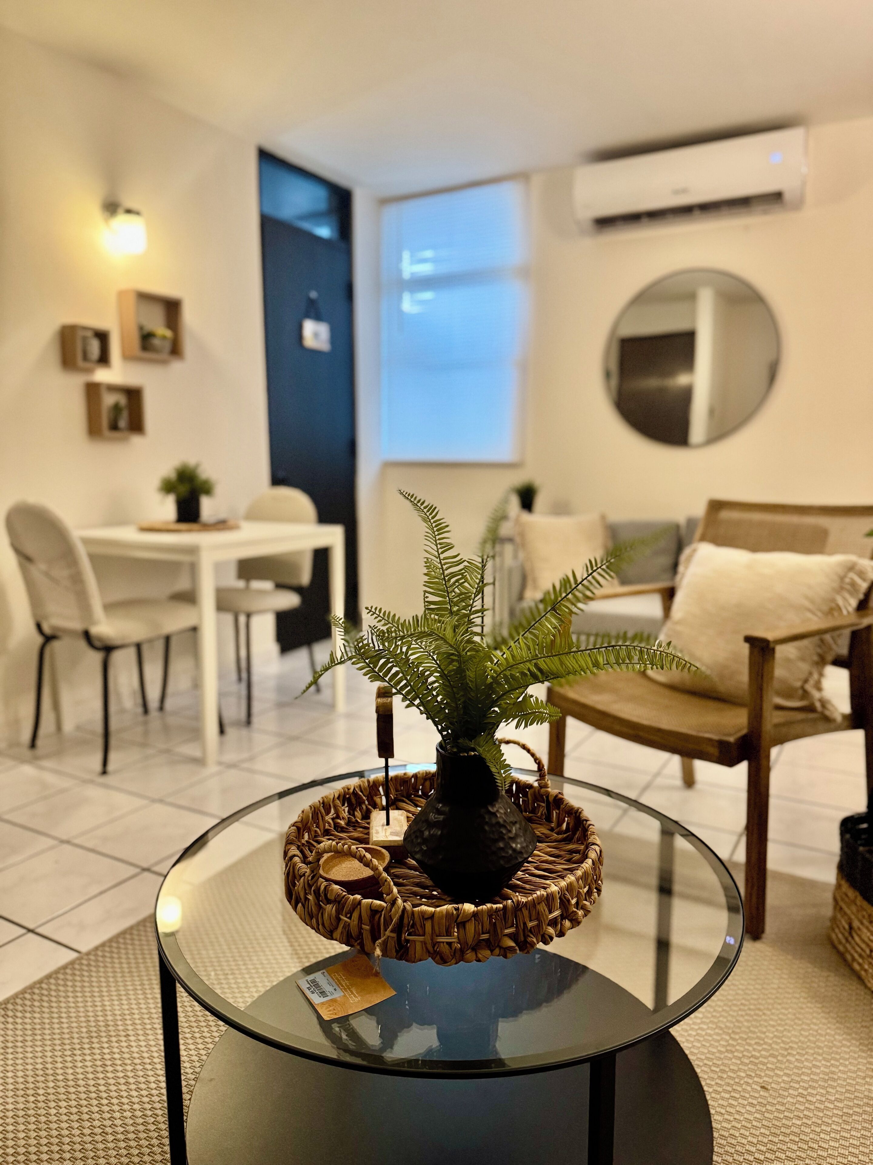 A modern living room with a glass coffee table, a dining area with a white table and chairs, a sofa with a cushion, and a large mirror on the wall.