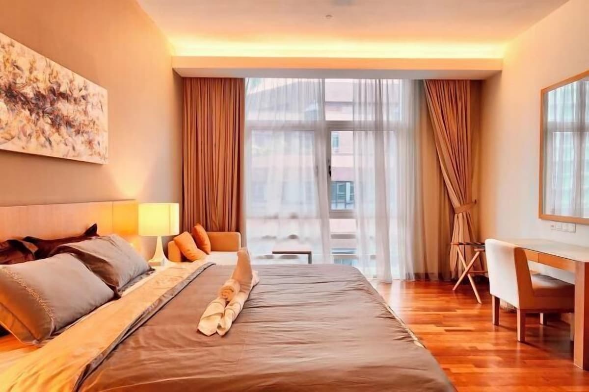 Habitación de hotel con cama, escritorio, silla, lámpara y un amplio ventanal con cortinas.