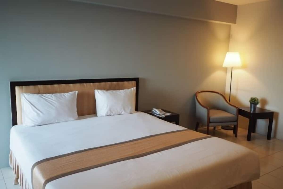 A hotel room with a large bed, a bedside table with a lamp, a chair, and a small table with a plant.