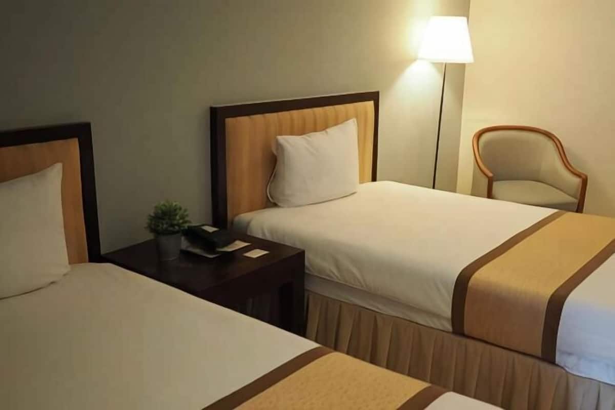 A hotel room with two beds, a nightstand with a lamp, and a chair.