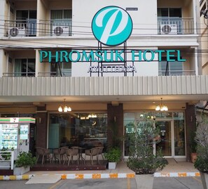 Un hotel con un logotipo destacado, un cartel con el nombre 'PHROMSUK HOTEL' y una entrada visible con mesas al aire libre.