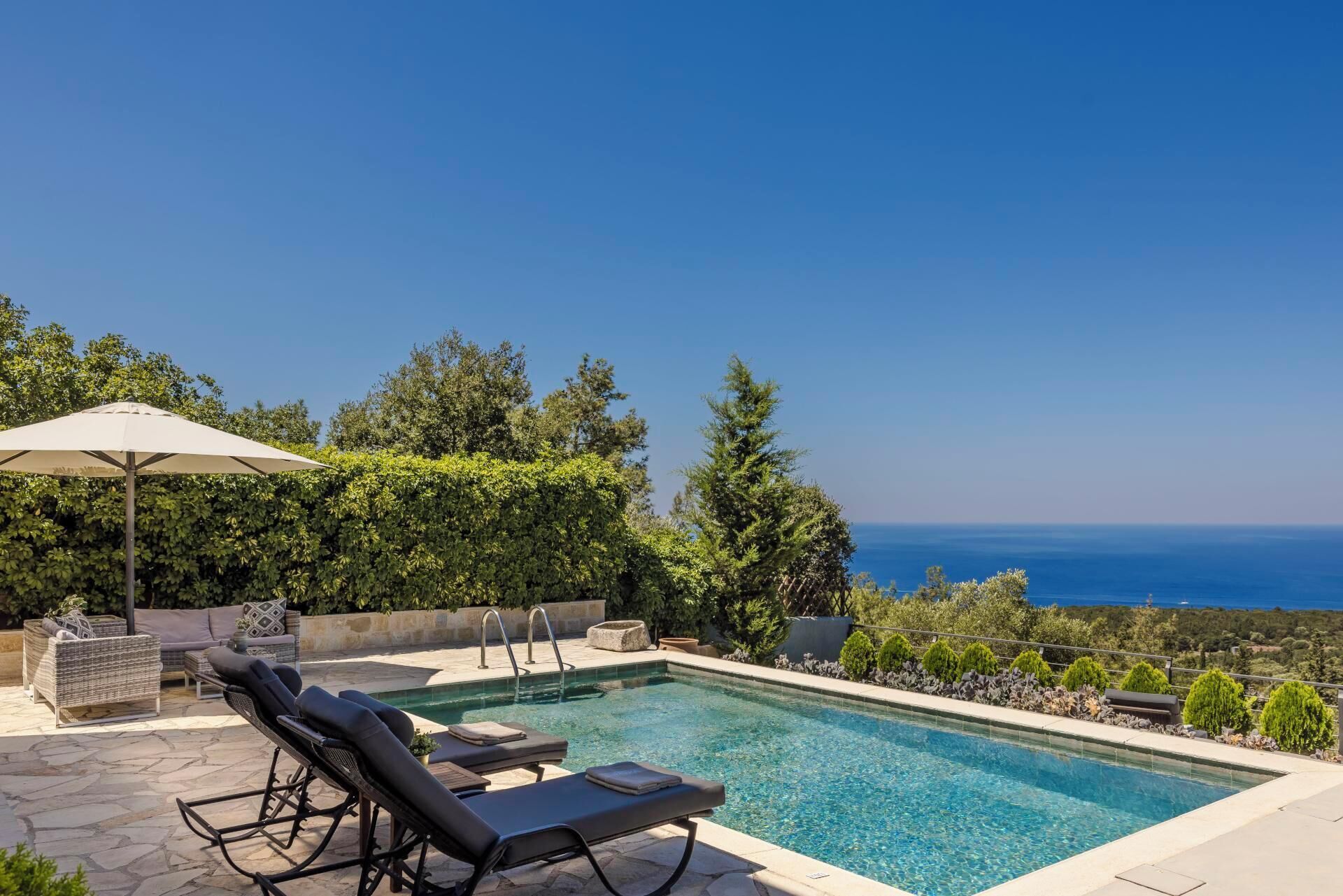 Área de piscina com espreguiçadeiras e um guarda-sol, cercada por vegetação e com vista para um corpo de água.