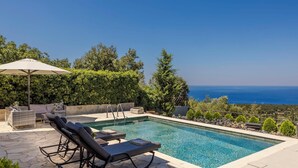 A pool area with lounge chairs and an umbrella, surrounded by greenery and overlooking a body of water. - Melanthi - Three Bedroom Villa, Sleeps 6 (Fiskardo)