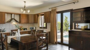 A modern kitchen with wooden cabinets, a dining table with chairs, a built-in oven, and a view of the outdoors. - Melanthi - Three Bedroom Villa, Sleeps 6 (Fiskardo)