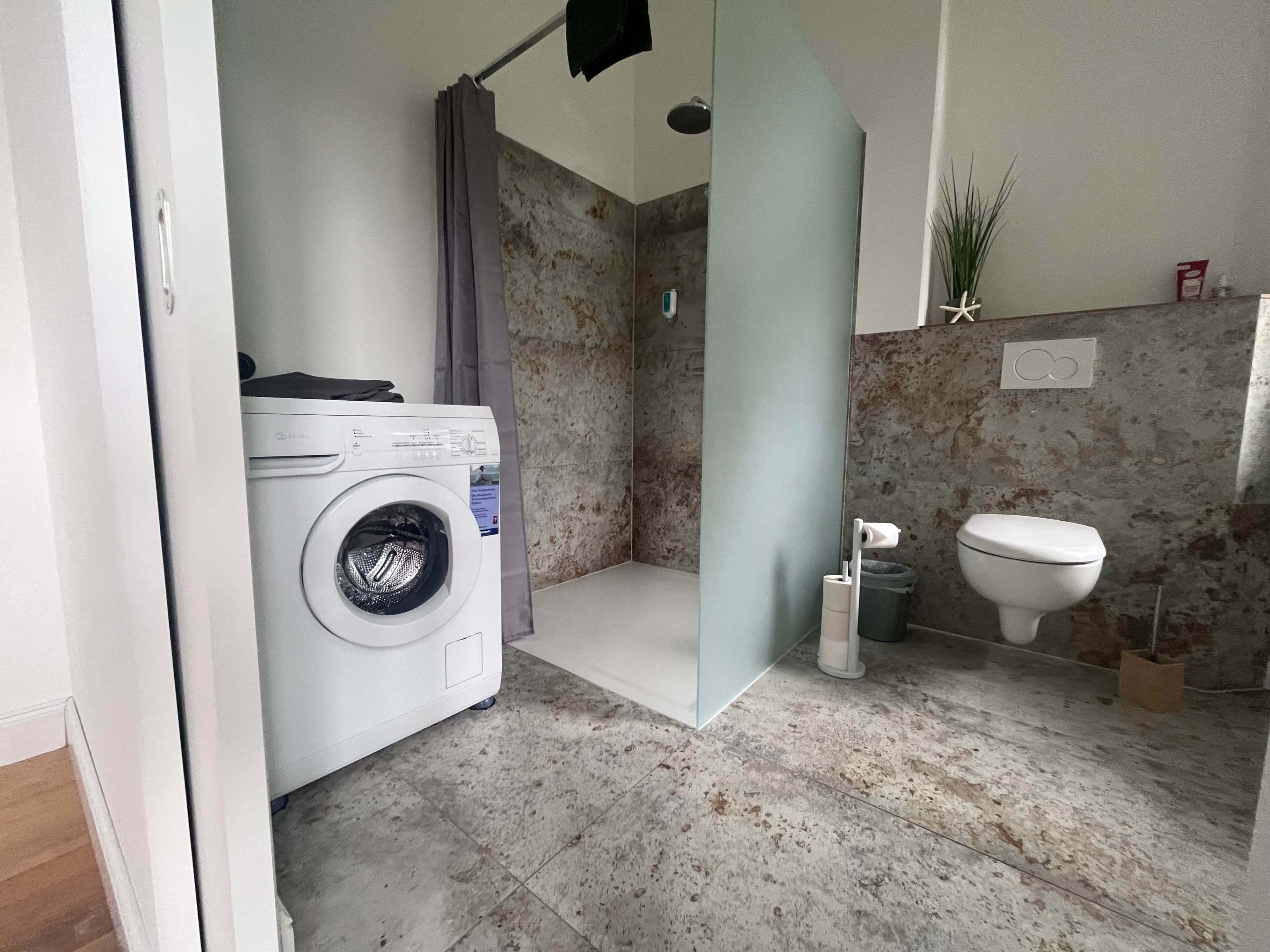 Et lille badeværelse med vaskemaskine, bruser, toilet og en lille skraldespand.