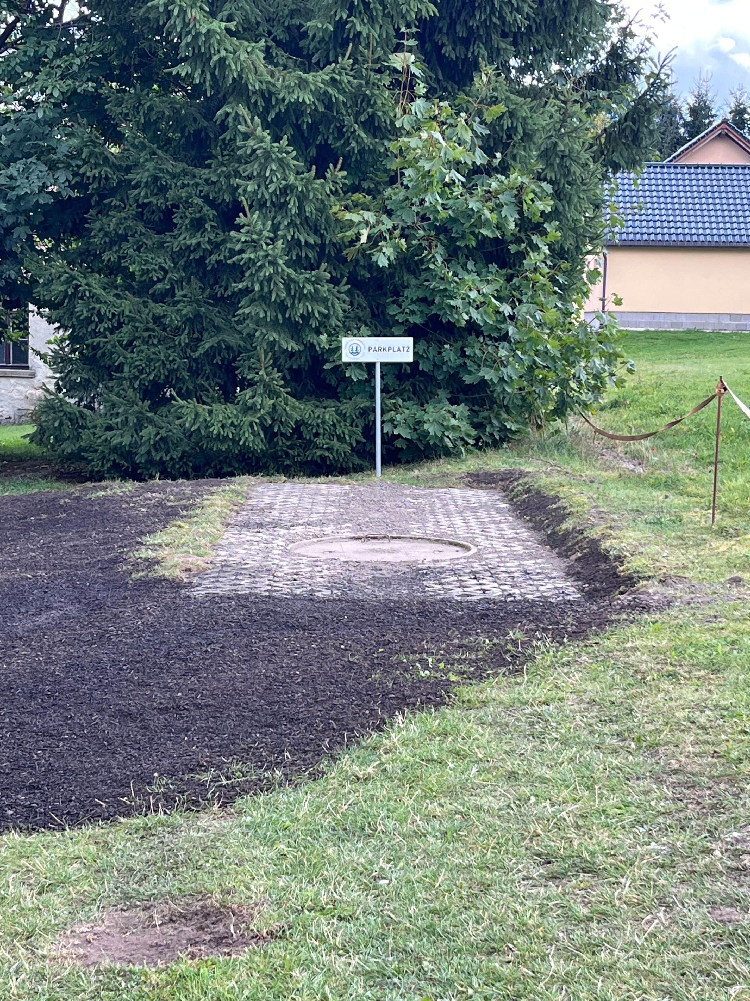 Een verhard gebied met een bord waarop 'Parkplatz' staat, omringd door groen en een gebouw met een blauw dak.