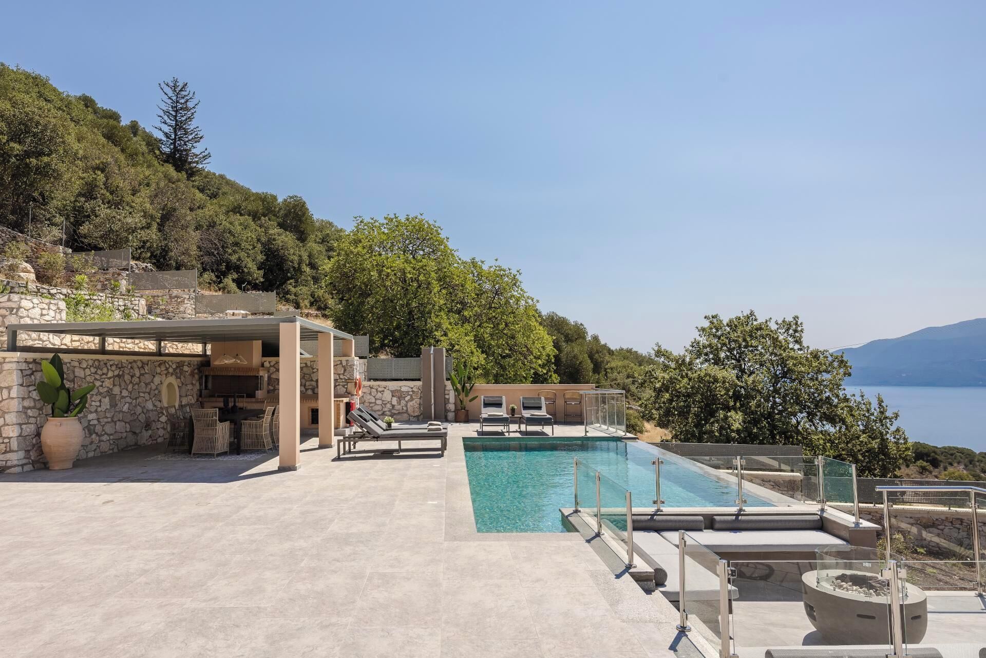 A pool area with lounge chairs and a stone wall, overlooking a body of water and hills.