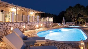 En pool med solstolar och en stenmur, omgiven av en pergola med belysning.