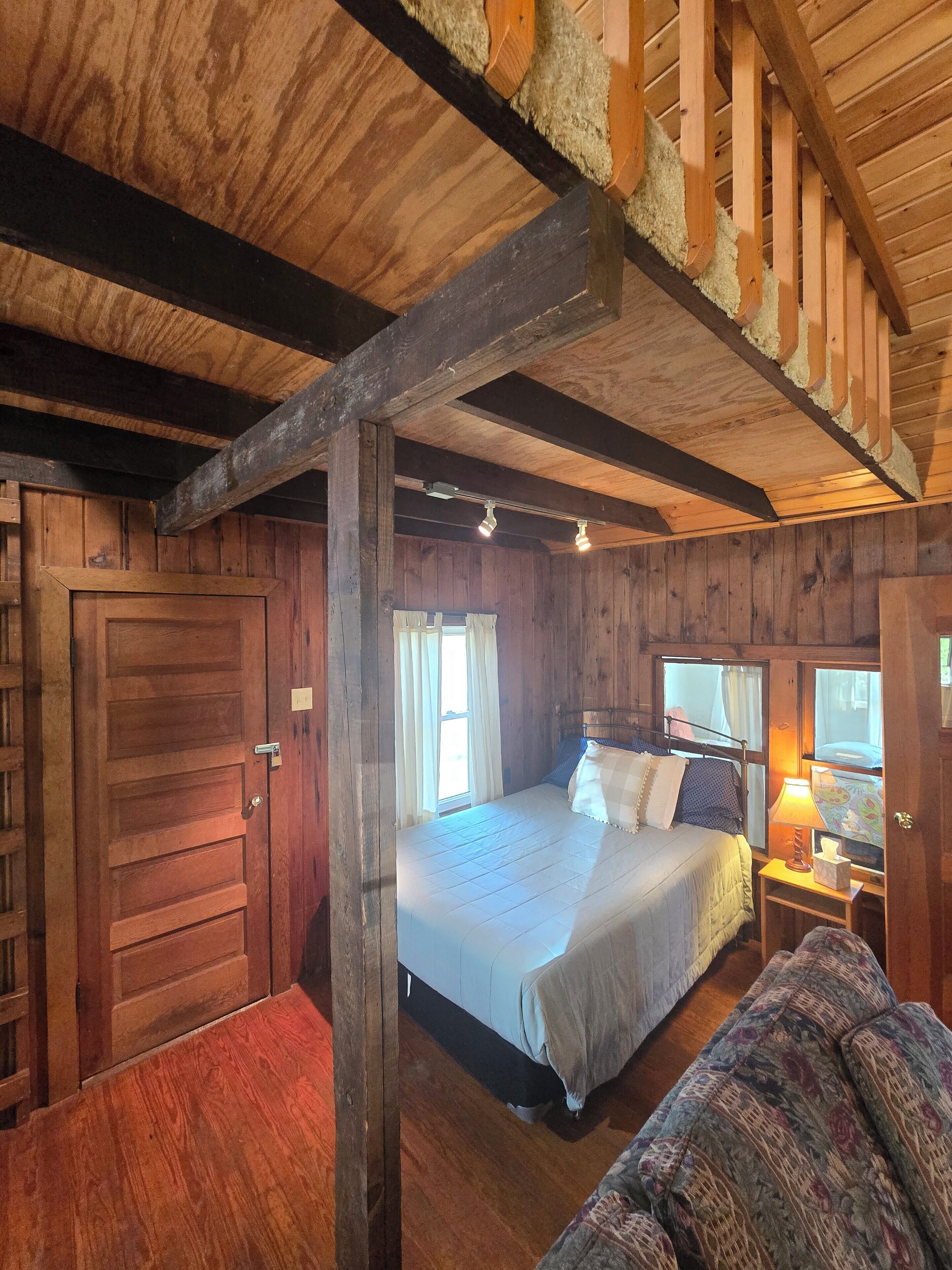 Een houten kamer in chaletstijl, met een bed, nachtkastje, raam met gordijnen en een deur.