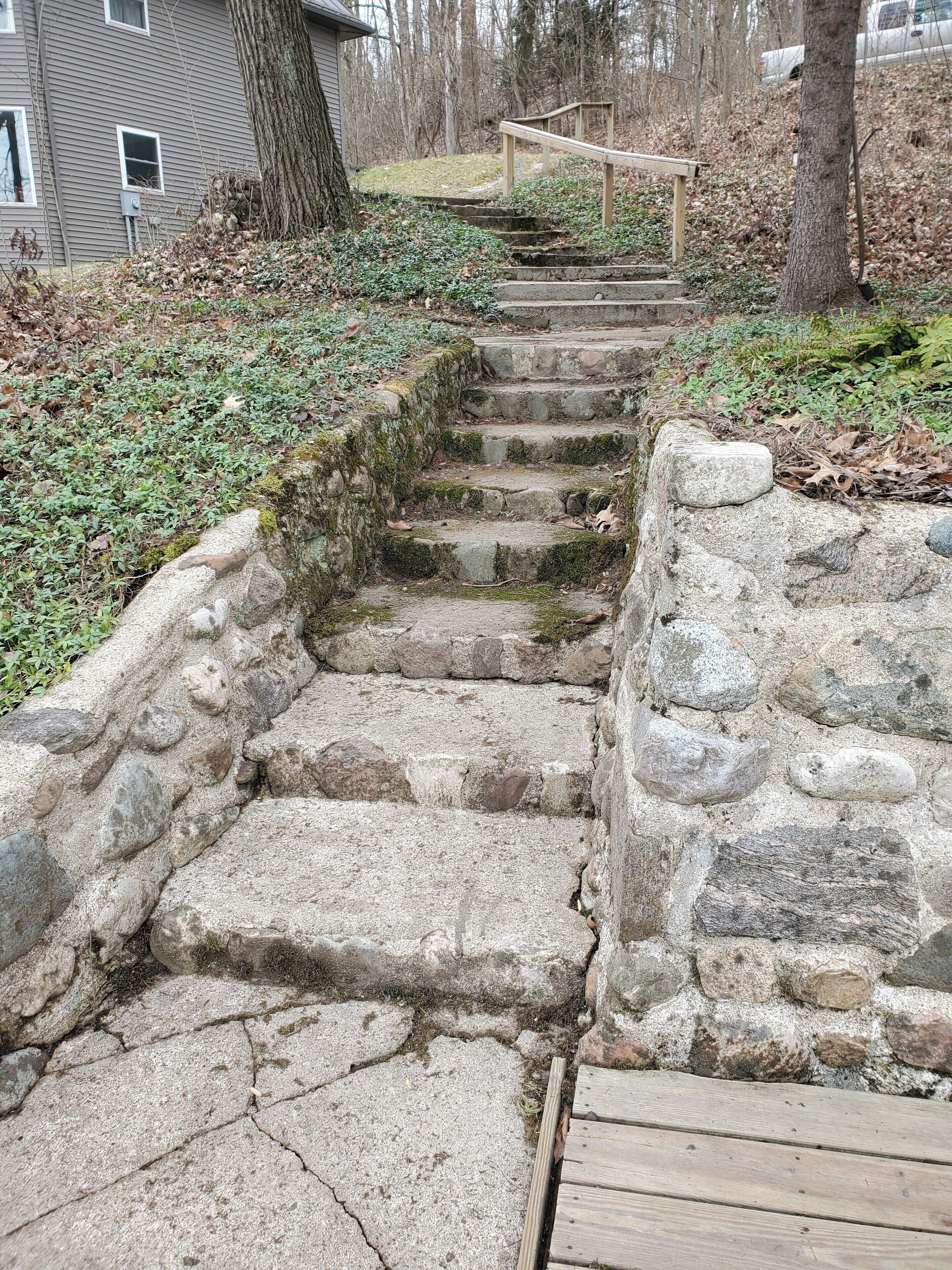 A set of stone steps leading up to a wooden deck, surrounded by a natural landscape with trees and a house in the background.