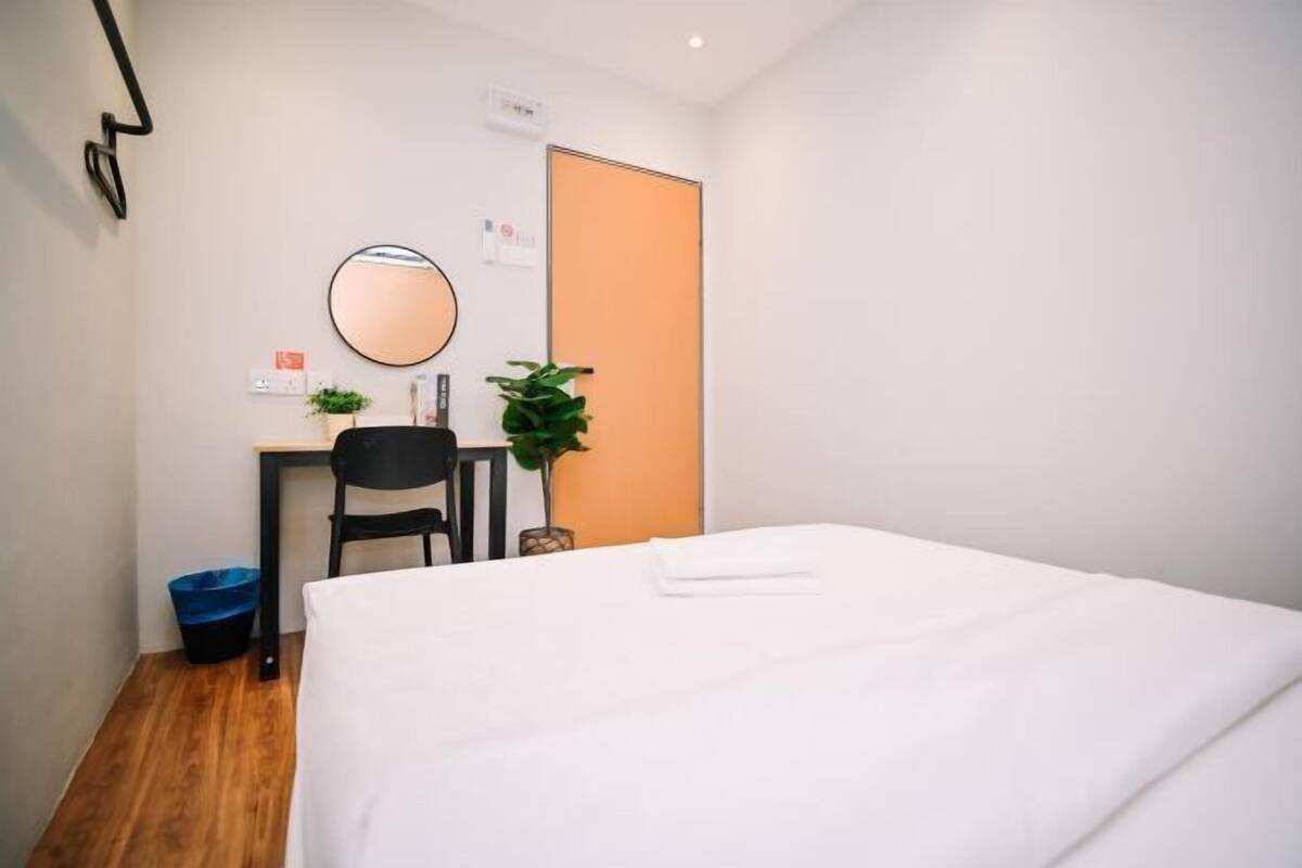 Hotellihuone, jossa on sänky, työpöytä tuolilla, pyöreä peili, ruukkukasvi ja sininen roskakori.