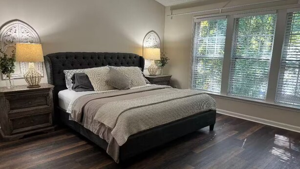 A bedroom with a dark upholstered bed, two bedside tables with lamps, a large window with blinds, and a wooden floor. - Beautiful and cozy house in Duluth! (Duluth)