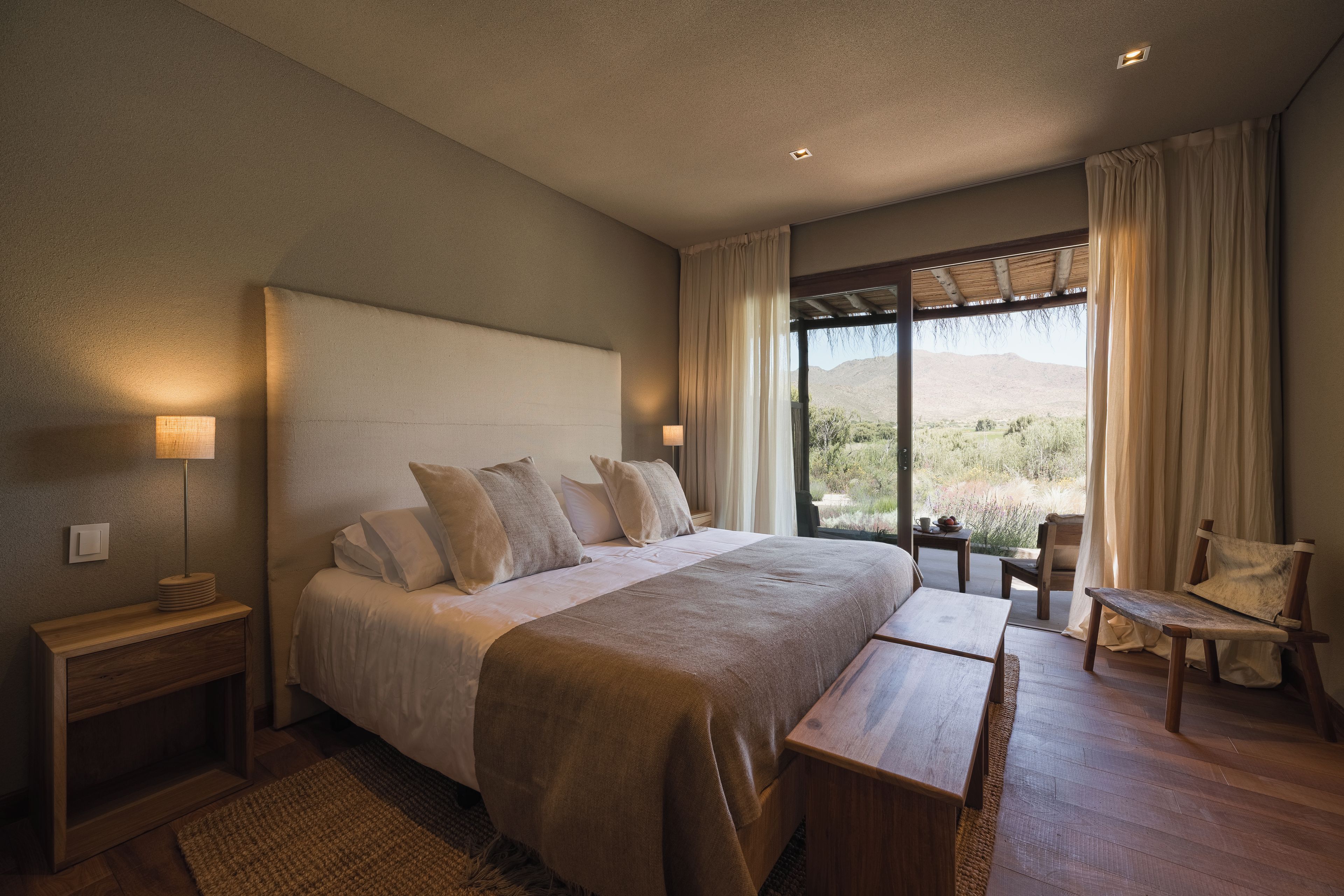 A bedroom with a large bed, bedside tables, a bench, a chair, and a view of the outdoors.