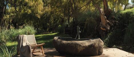 A wooden deck with a stone basin and a wicker chair, surrounded by greenery.