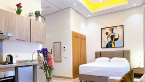 Ein modernes Hotelzimmer mit einer Küche, einem Bett, einem Nachttisch mit Blumen und einem Bild an der Wand.