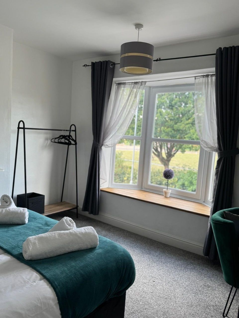 A bedroom with a bed, a window with curtains, a hanging light fixture, a small table with a potted plant, and a chair.