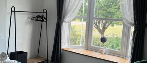 A bedroom with a bed, a window with curtains, a hanging light fixture, a small table with a potted plant, and a chair.