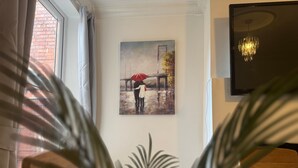 A room with a painting of a couple under an umbrella, a chandelier, and a plant in the foreground. - Station View | Family | Parking | WIFI | Sleeps 6 (Leicestershire)