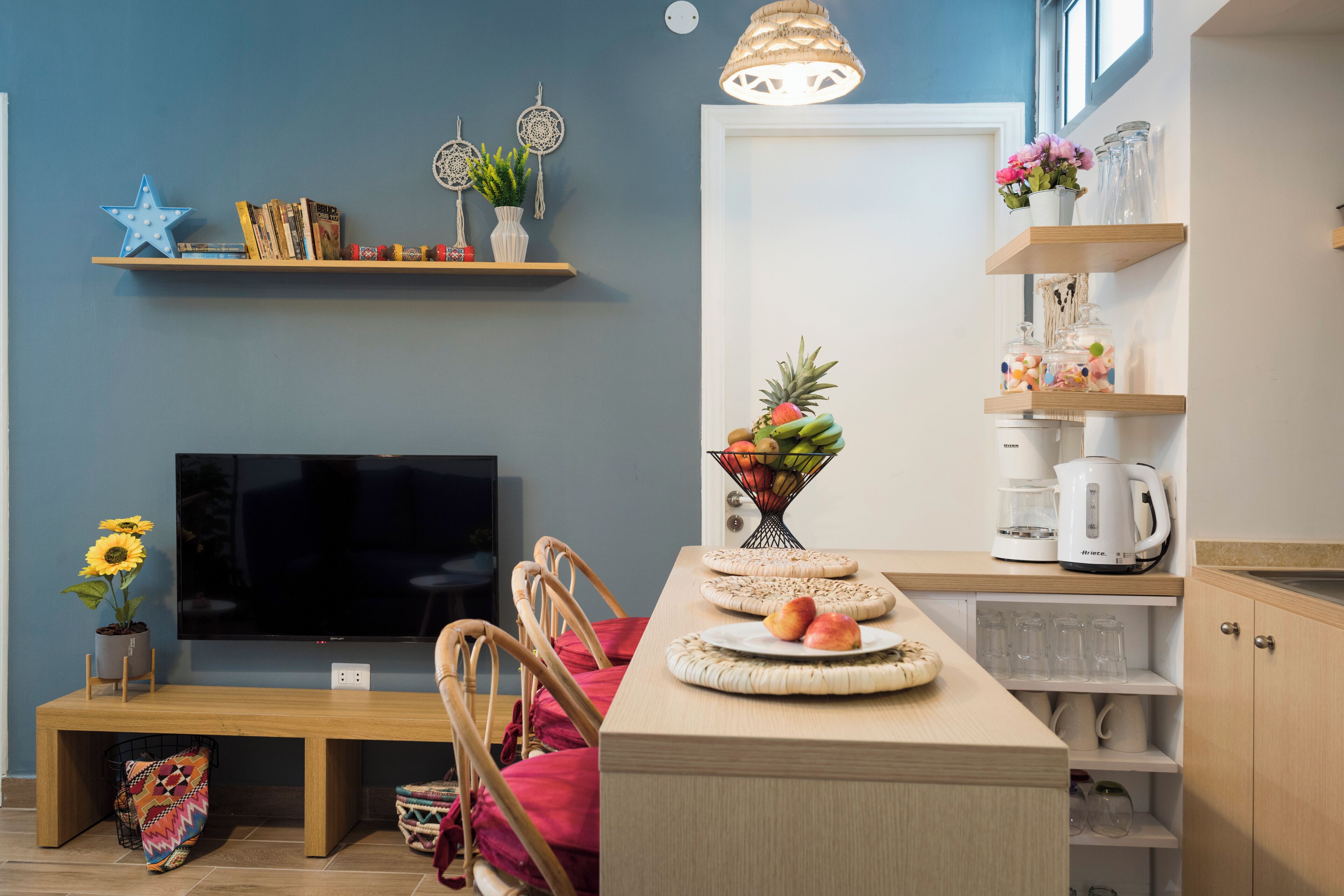 Una sala de estar moderna con televisor de pantalla plana, muebles de madera, una mesa de comedor con fruteros y una cocina con estantes y cafetera.