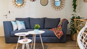 A modern living room with a grey sofa, a small round table with a vase of flowers, and a wicker chair. - Boho-Chic Cozy 2BR - Apt & Balcony in Mar Mikhael (Beirut)