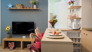A modern living room with a flat-screen TV, a dining table with chairs, a coffee table with decorative items, and a kitchen area with shelves and a coffee maker. - Boho-Chic Cozy 2BR - Apt & Balcony in Mar Mikhael (Beirut)