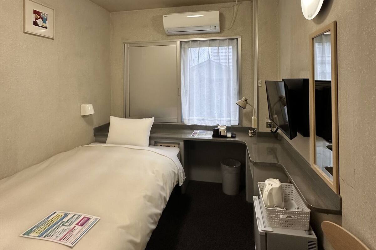 Habitación de hotel con cama, escritorio, televisor, unidad de aire acondicionado y ventana con cortinas.