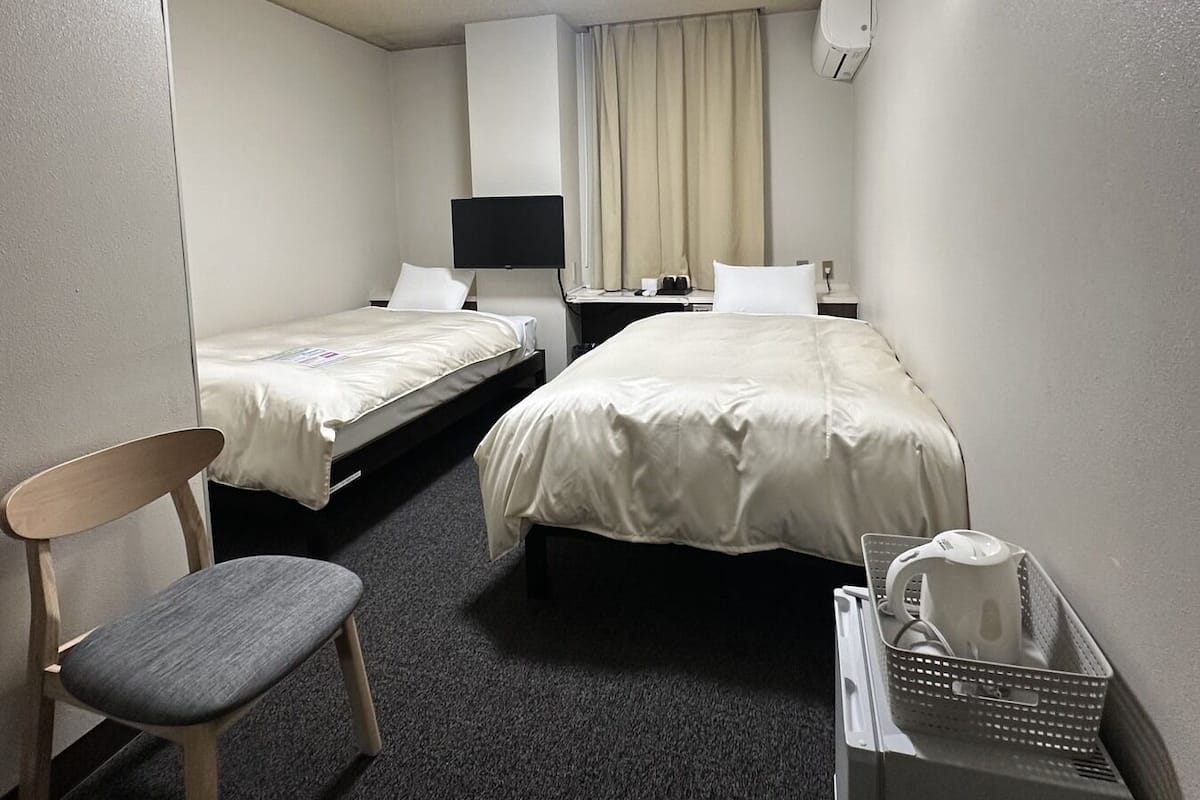 Habitación de hotel con dos camas, un escritorio, una silla, una lámpara de escritorio, aire acondicionado de pared y un cubo de basura.