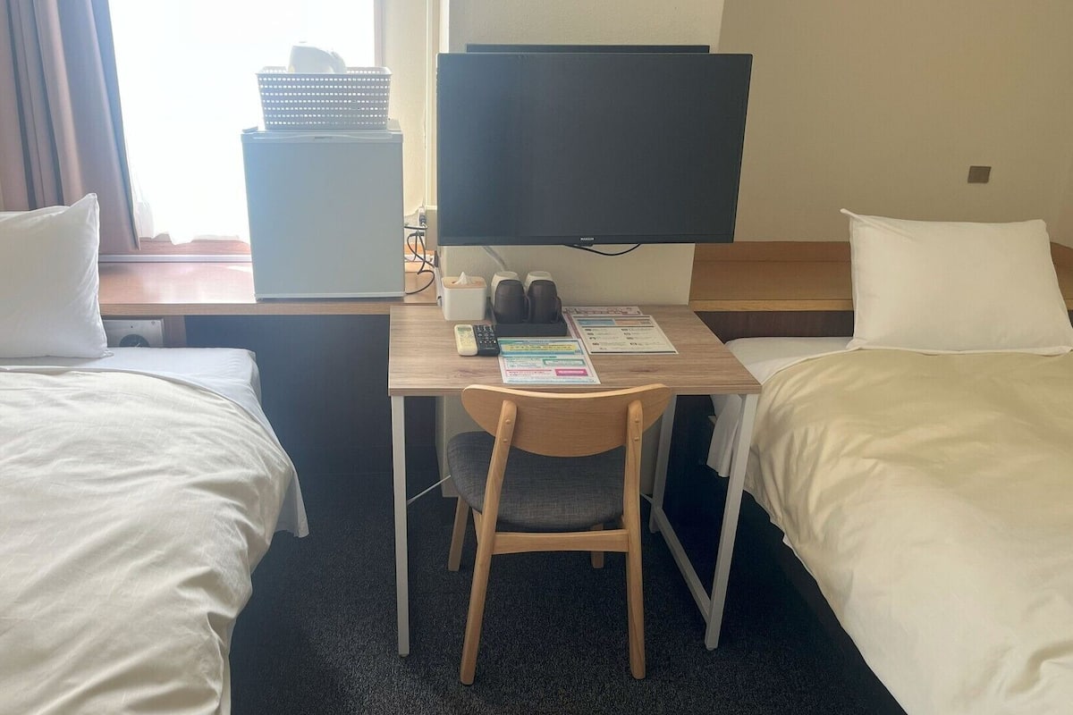 Habitación de hotel con dos camas individuales, un escritorio con una silla, un televisor y una mesita con una cesta y una caja de pañuelos.