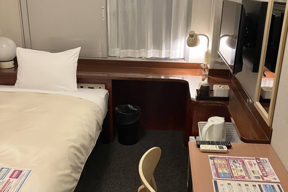 Habitación de hotel con una cama, un escritorio con silla, un televisor, una ventana con cortinas y un bote de basura.