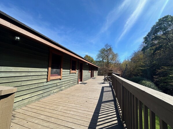A wooden deck with a railing, a building with multiple windows, and a view of trees and hills in the background.