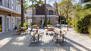 Een terras met meerdere houten tafels en stoelen, omringd door potplanten en bomen.