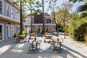 Un espace extérieur aménagé en terrasse, avec plusieurs tables et chaises en bois, entouré de plantes en pot et d’arbres.