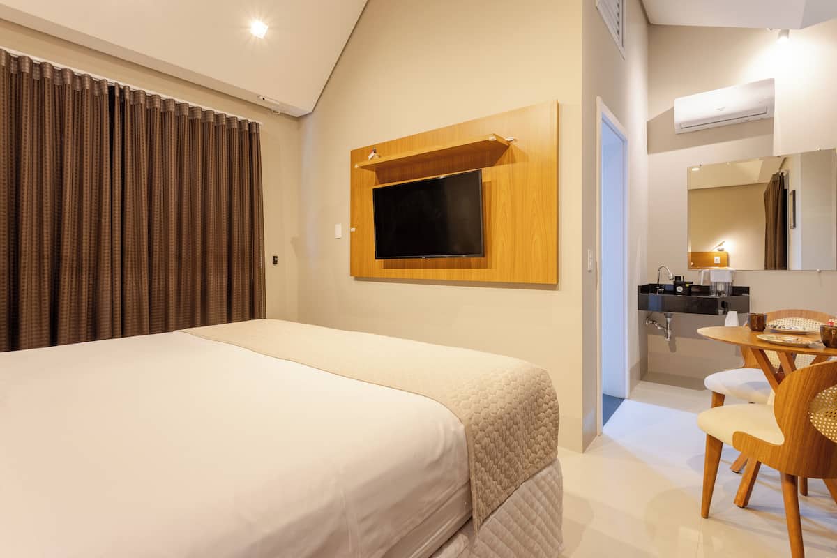 A hotel room with a large bed, a TV mounted on the wall, a dining area with a table and chairs, and a bathroom visible through an open door.