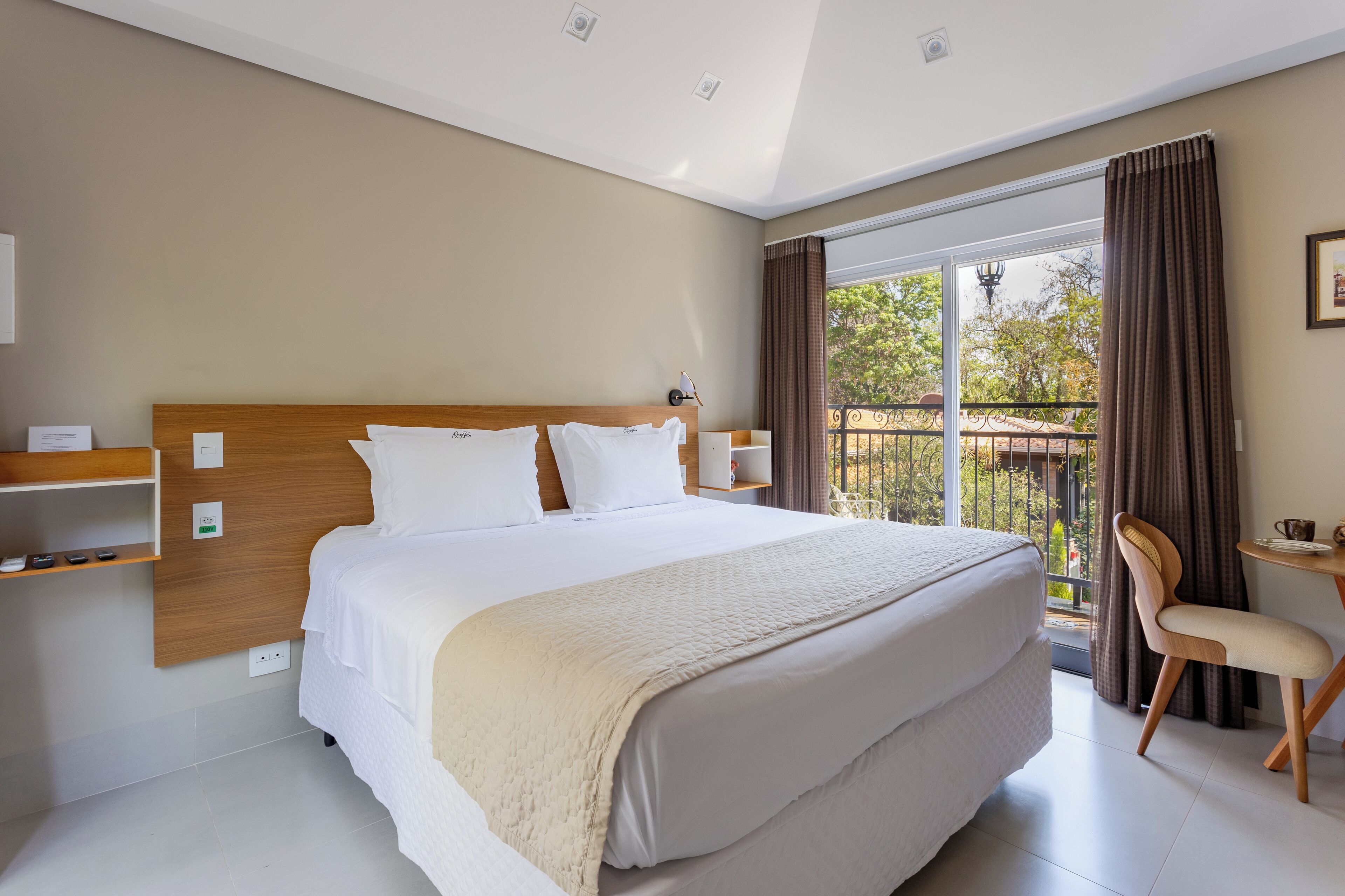 A hotel room with a large bed, a wooden headboard, a bedside table, a chair, and a balcony with curtains.