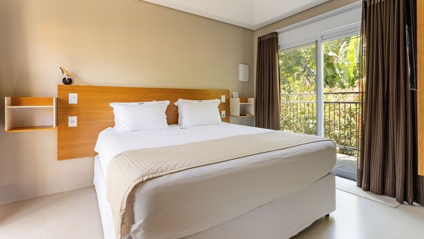 Een moderne hotelkamer met een groot bed, een houten hoofdbord, een nachtkastje en uitzicht op groen buiten het raam.