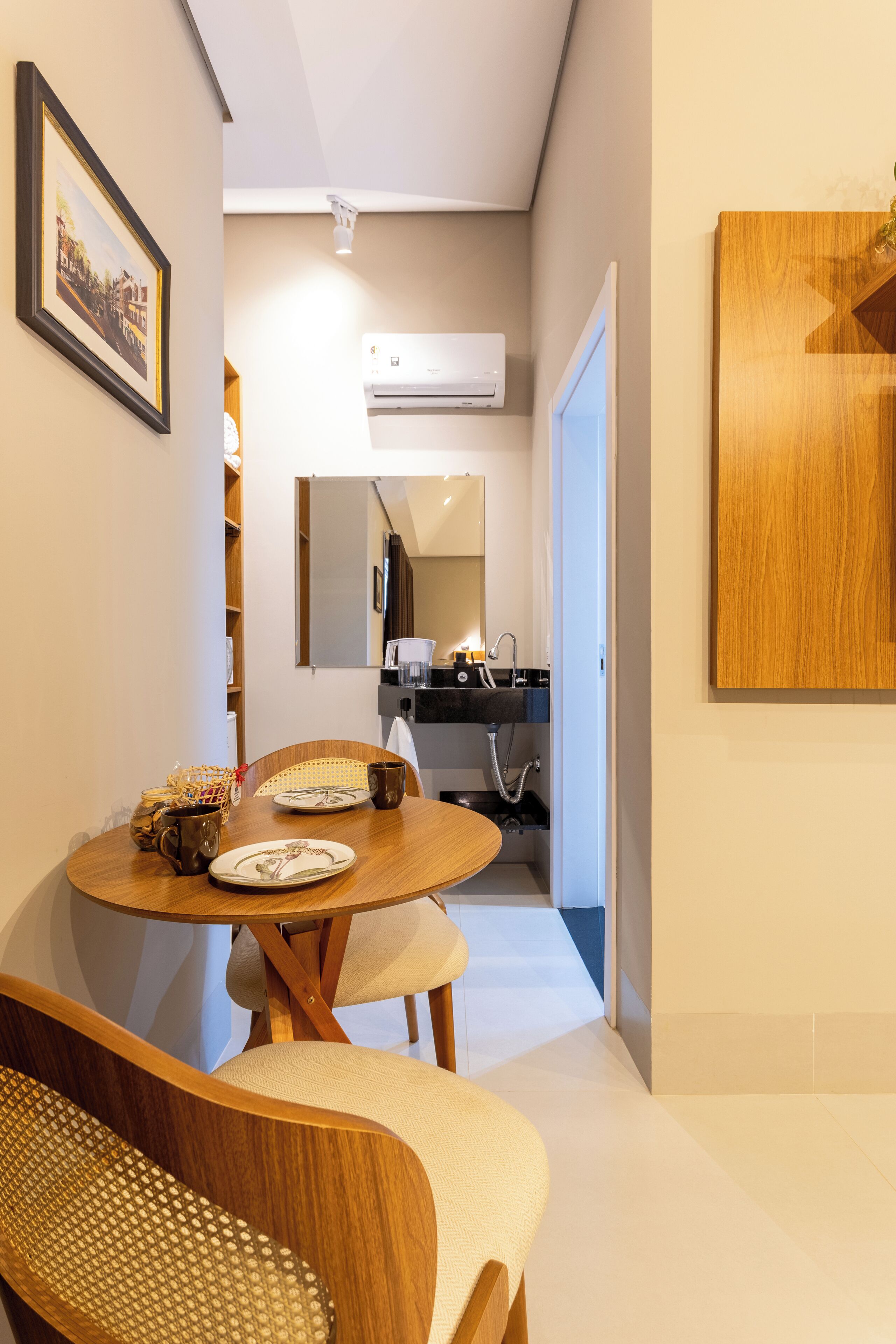 A compact dining area with a round table, chairs, and a bench. A mirror and an air conditioning unit are mounted on the wall.