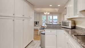 A kitchen with white cabinets, a granite countertop, a built-in microwave, and a view to the outside. - Short Easy Flat Walk to Beach - Pelican Watch 8a - Lakefront Villa w/ Pool (Pawleys Island)